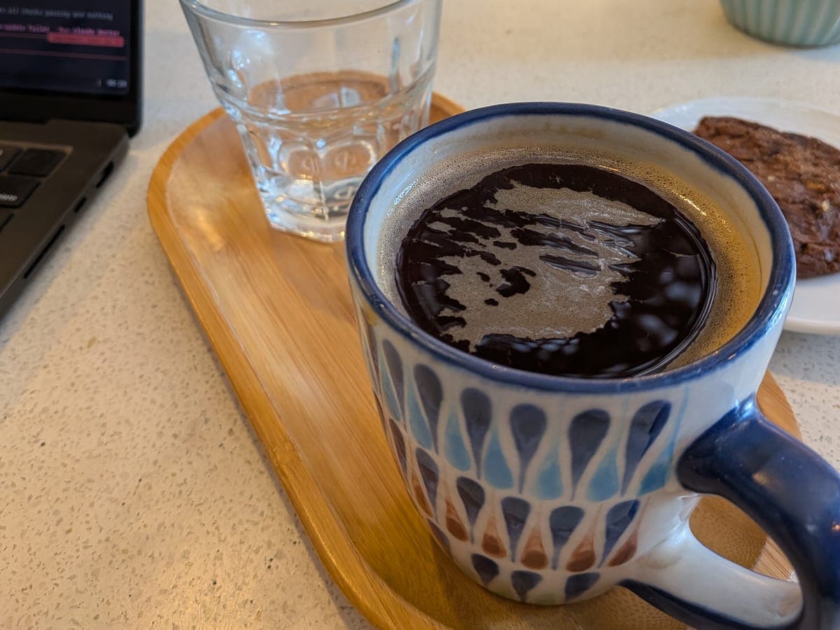 Taza de café negro y un vaso de agua sobre una bandeja de madera, en la mesa de una cafetería.
